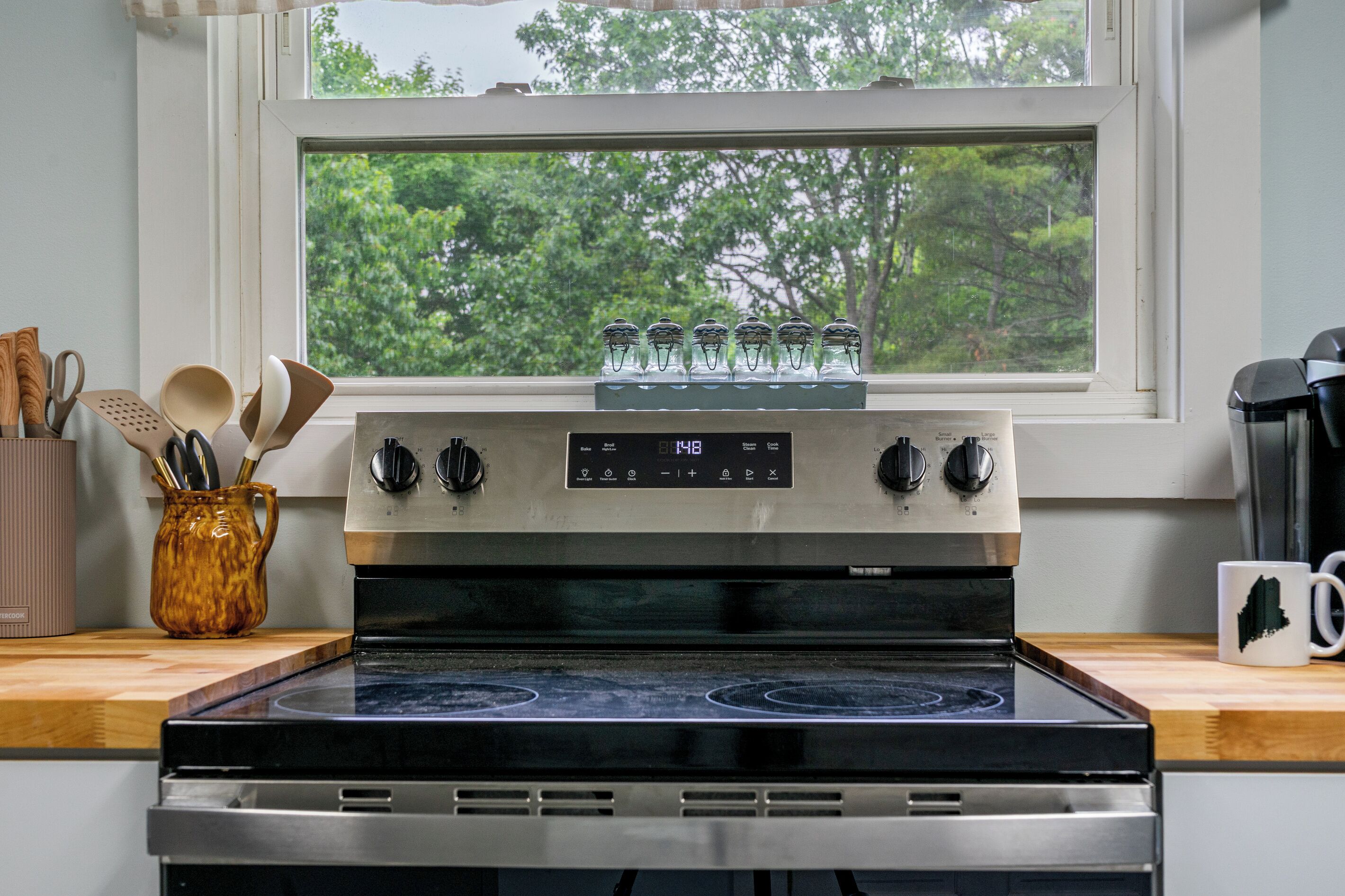 Une cuisine équipée d’une cuisinière, d’une fenêtre donnant sur des arbres et d’ustensiles de cuisine posés sur le plan de travail.