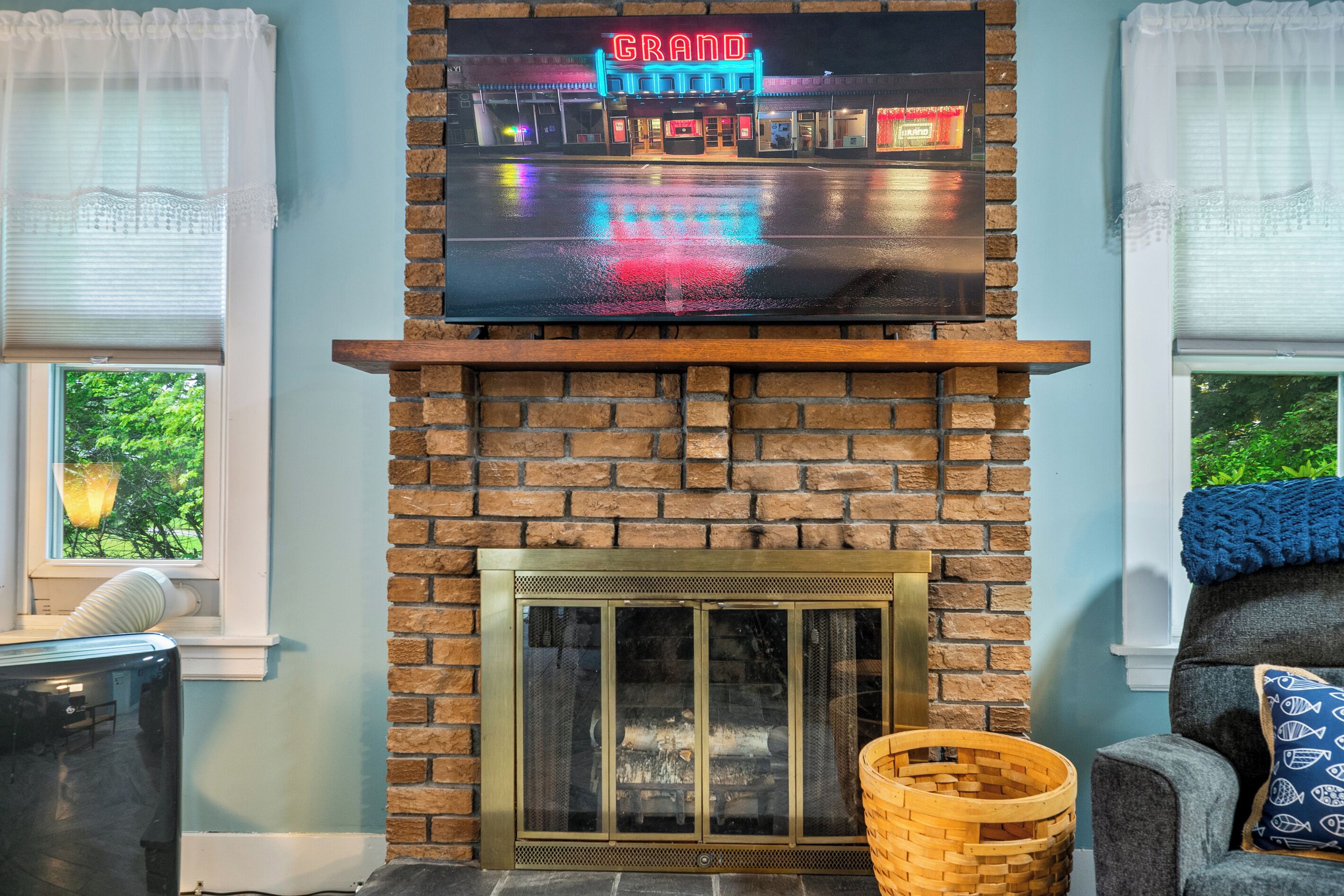 A living room with a brick fireplace, a mounted TV displaying a neon sign, and a window with a view of greenery.