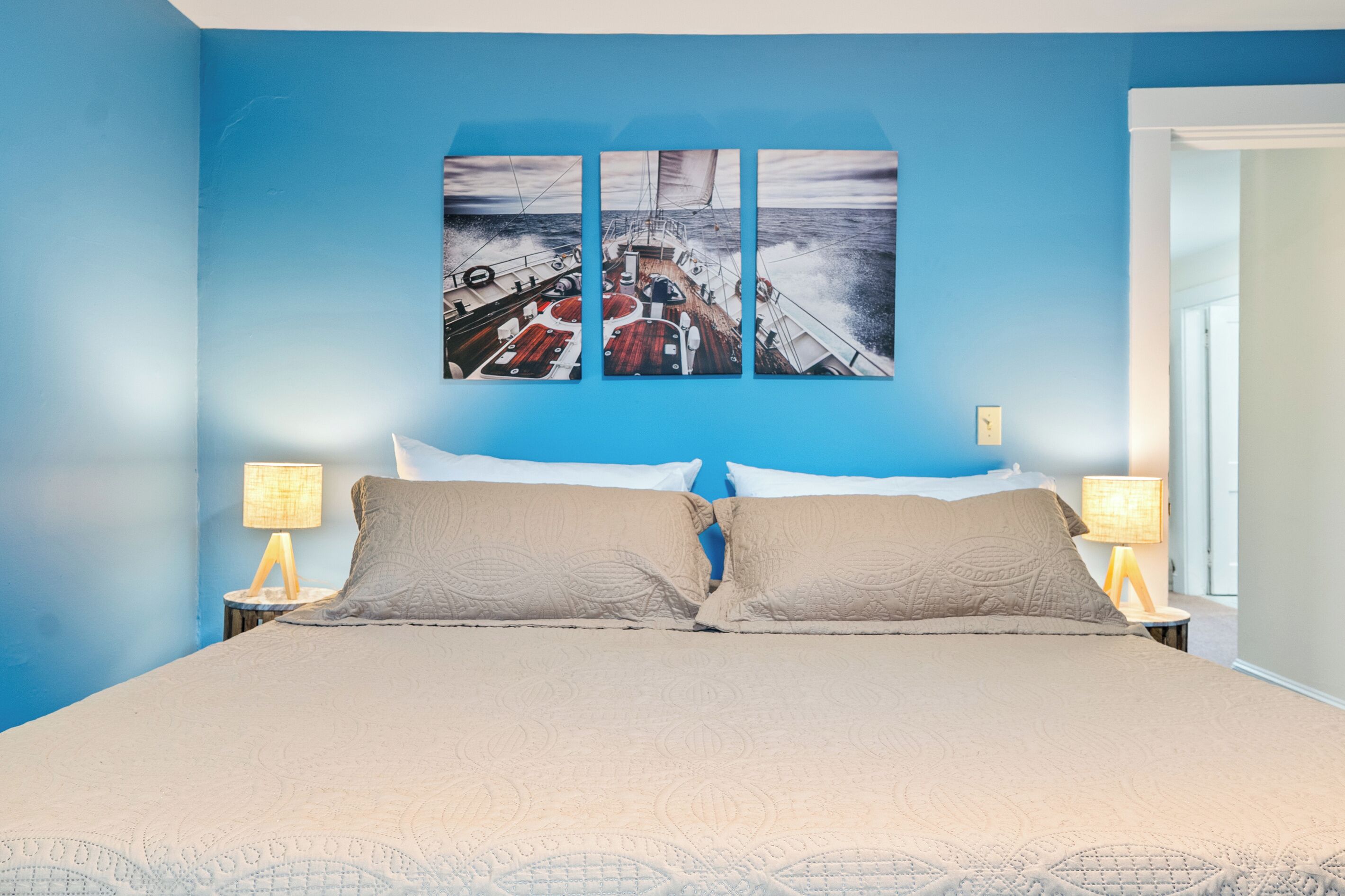 Un lit recouvert d’une couvre-lit beige, avec deux oreillers et un tableau représentant un voilier accroché au mur.