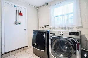 A laundry room with two washing machines, a door, and a window with a curtain.