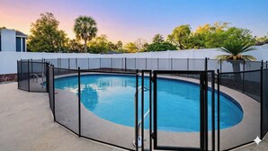 Une piscine entourée d’une clôture de sécurité, d’un mur blanc et d’arbres.