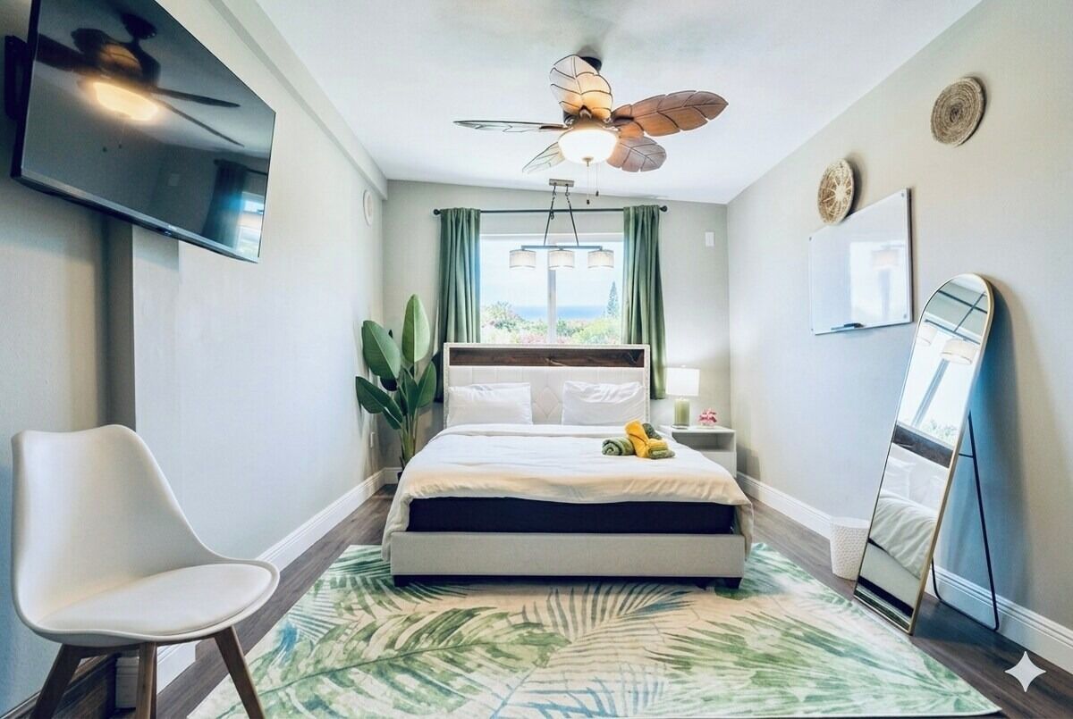 A bedroom with a bed, a chair, a ceiling fan, a TV, and a window with curtains.