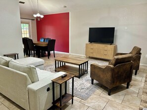 Ein Wohnzimmer mit Sofa, Sessel, Couchtisch, Fernseher und einem Essbereich mit Stühlen und Tisch.