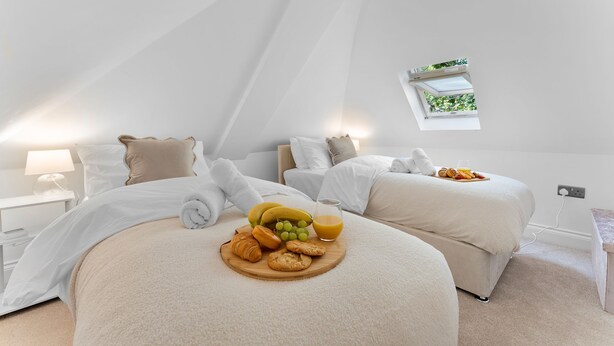 Ein Schlafzimmer mit zwei Betten, einem Frühstückswagen mit Croissants, Trauben und Orangensaft, sowie einem kleinen Fenster mit Blick auf Grünflächen.