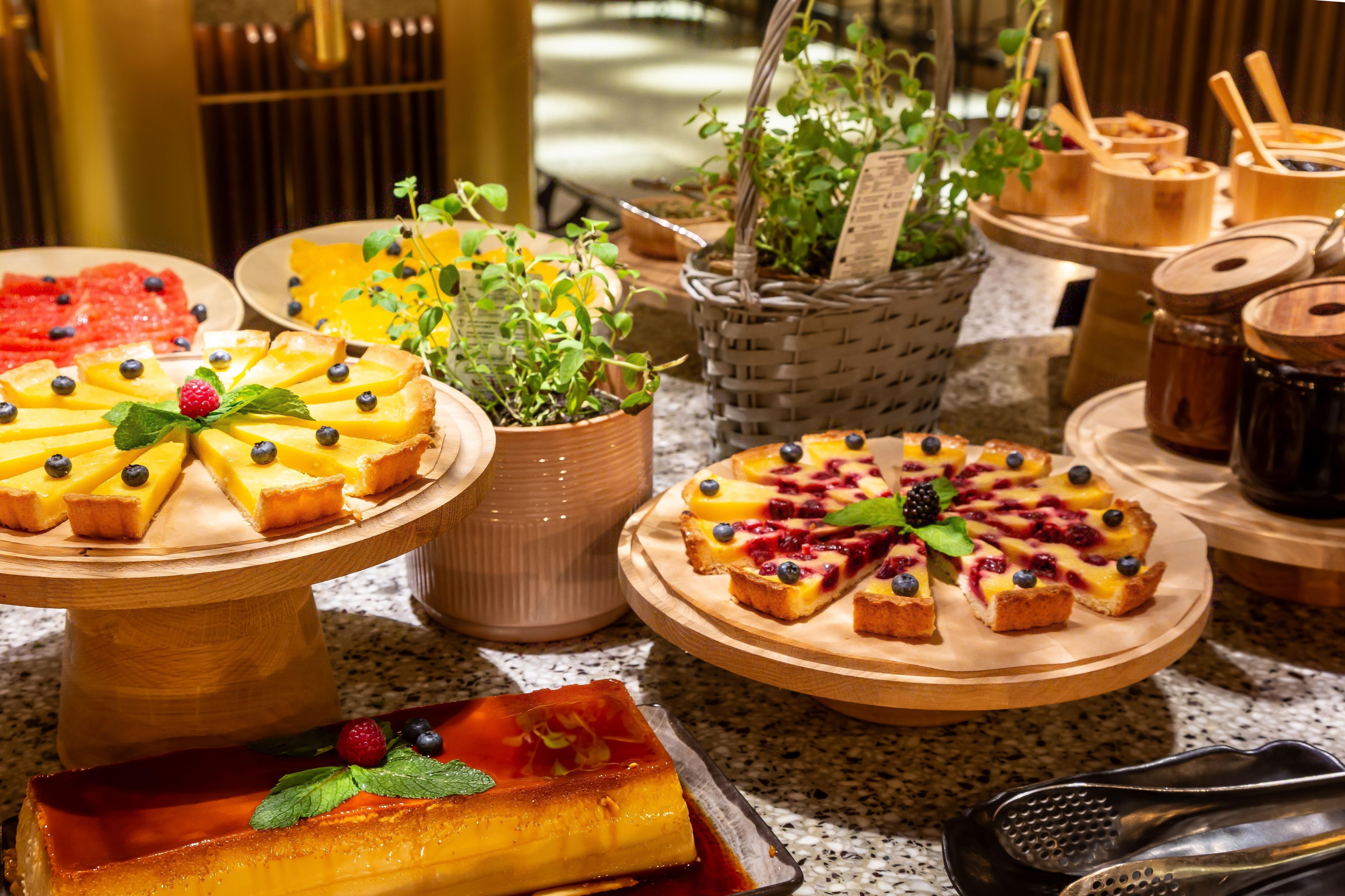Variedad de postres en bandejas de madera, incluyendo flan y tartaletas con frutas, con un fondo de utensilios de cocina y una planta en maceta.