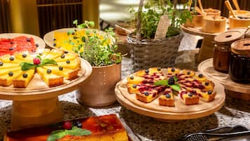 Variedad de postres en bandejas de madera, incluyendo flan y tartaletas con frutas, con un fondo de utensilios de cocina y una planta en maceta.