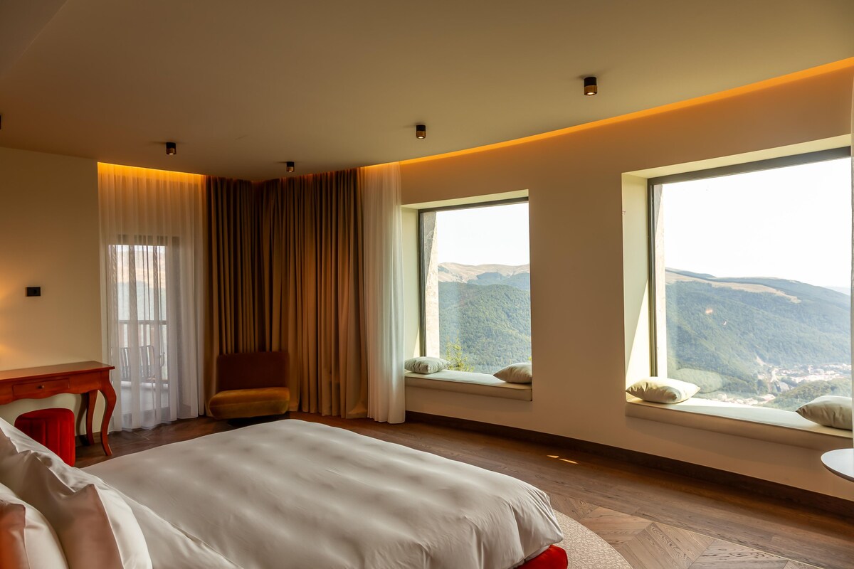 A hotel room with a large bed, a desk, and a chair. There are two windows with mountain views.