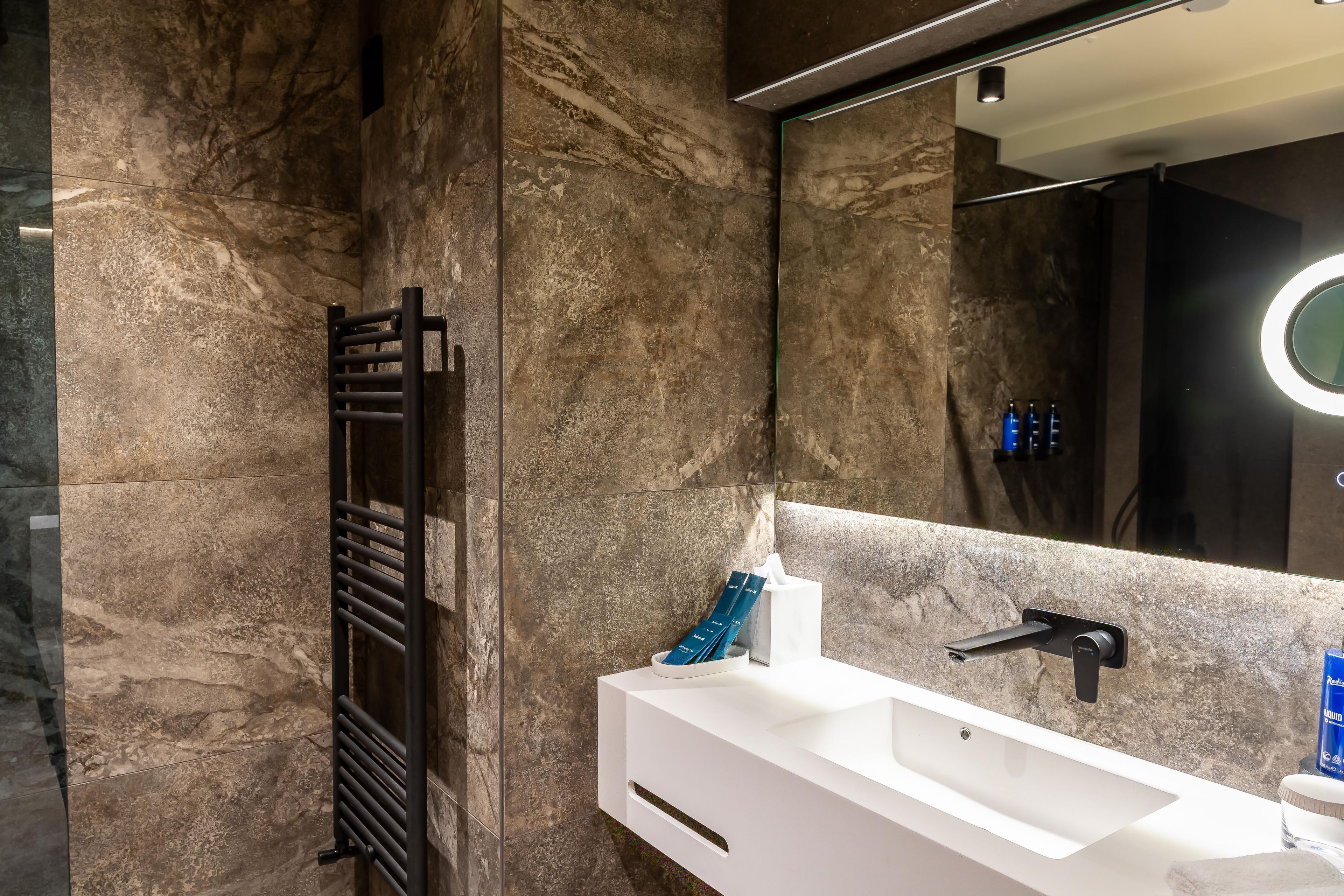 A modern bathroom with a marble wall, a black towel rack, a white sink, and a mirror with a circular light.