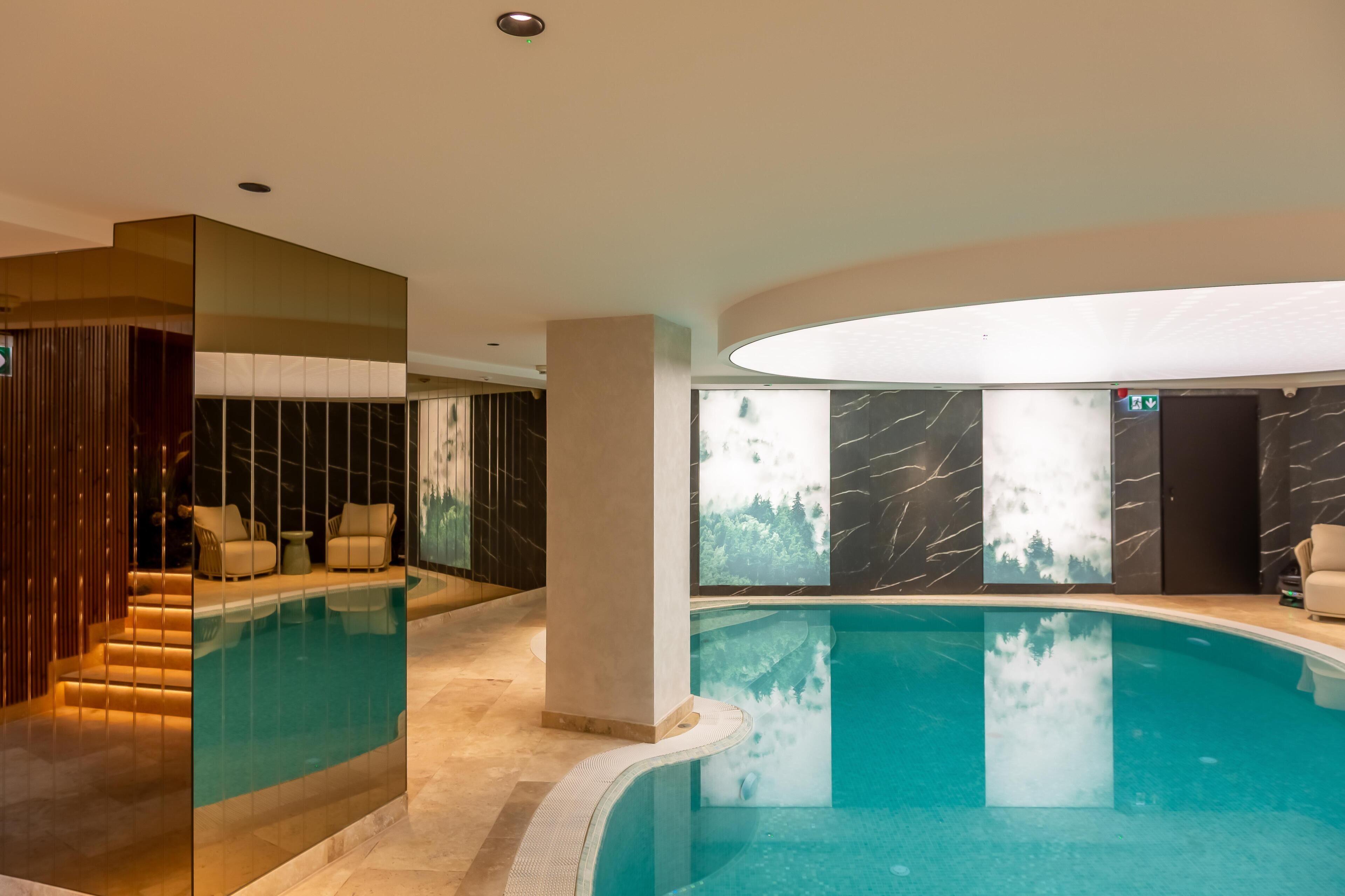 Piscina interior moderna con pared de vidrio, zona de estar y vistas a un bosque a través de las ventanas.