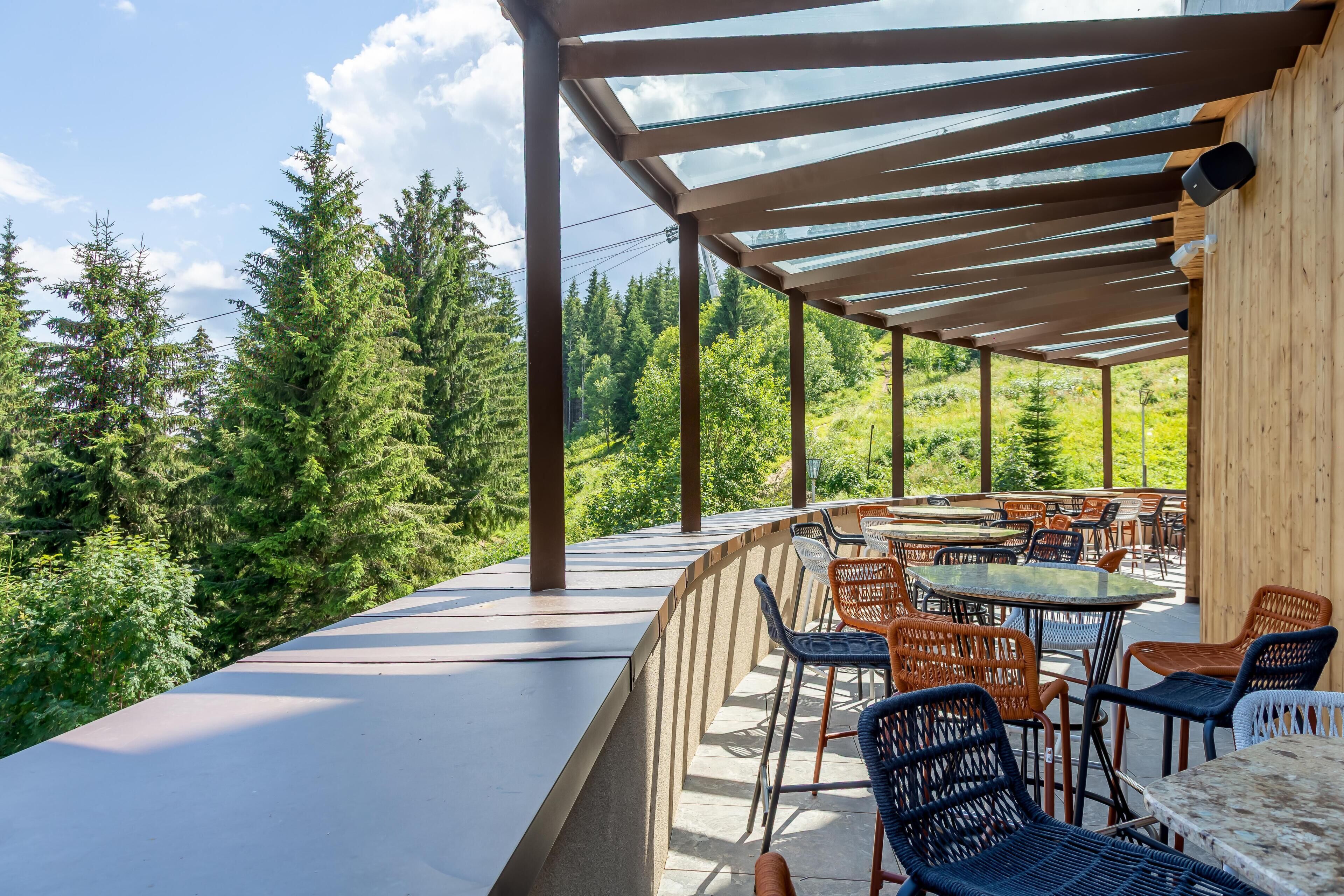 An outdoor patio with tables and chairs, surrounded by a wooden pergola and a view of a forest.