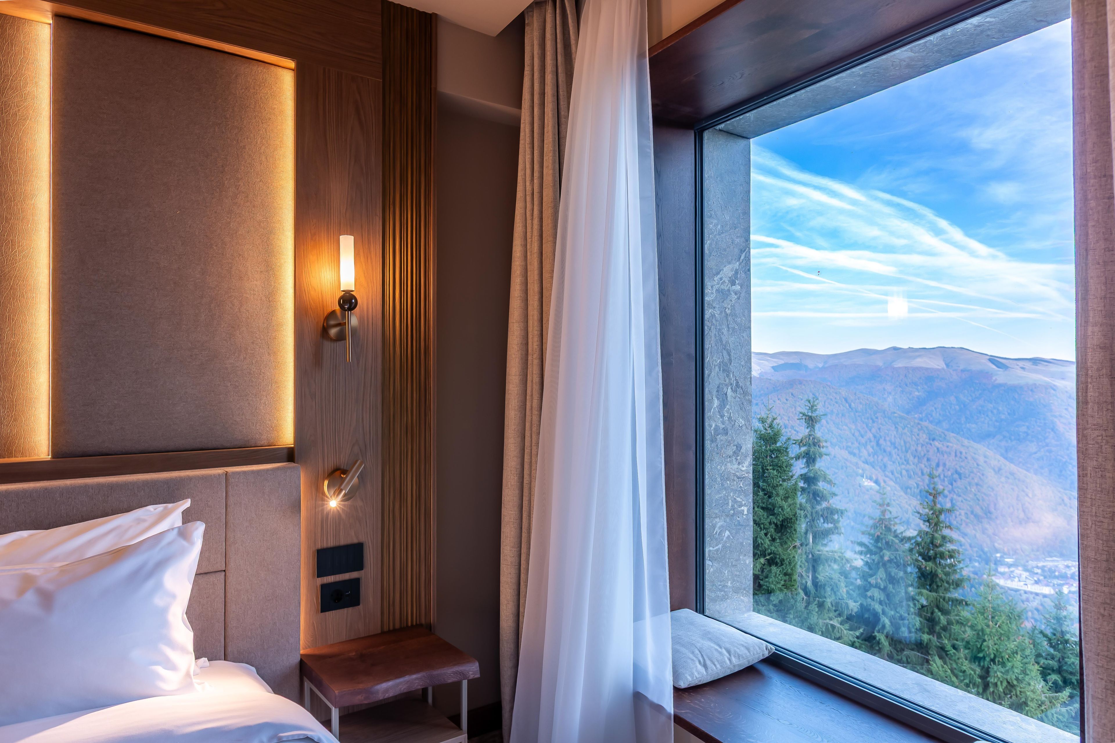 A hotel room with a bed, a bedside table, a nightstand, a wall-mounted lamp, and a large window with a mountain view.