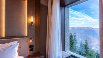 A hotel room with a bed, a bedside table, a nightstand, a wall-mounted lamp, and a large window with a mountain view.
