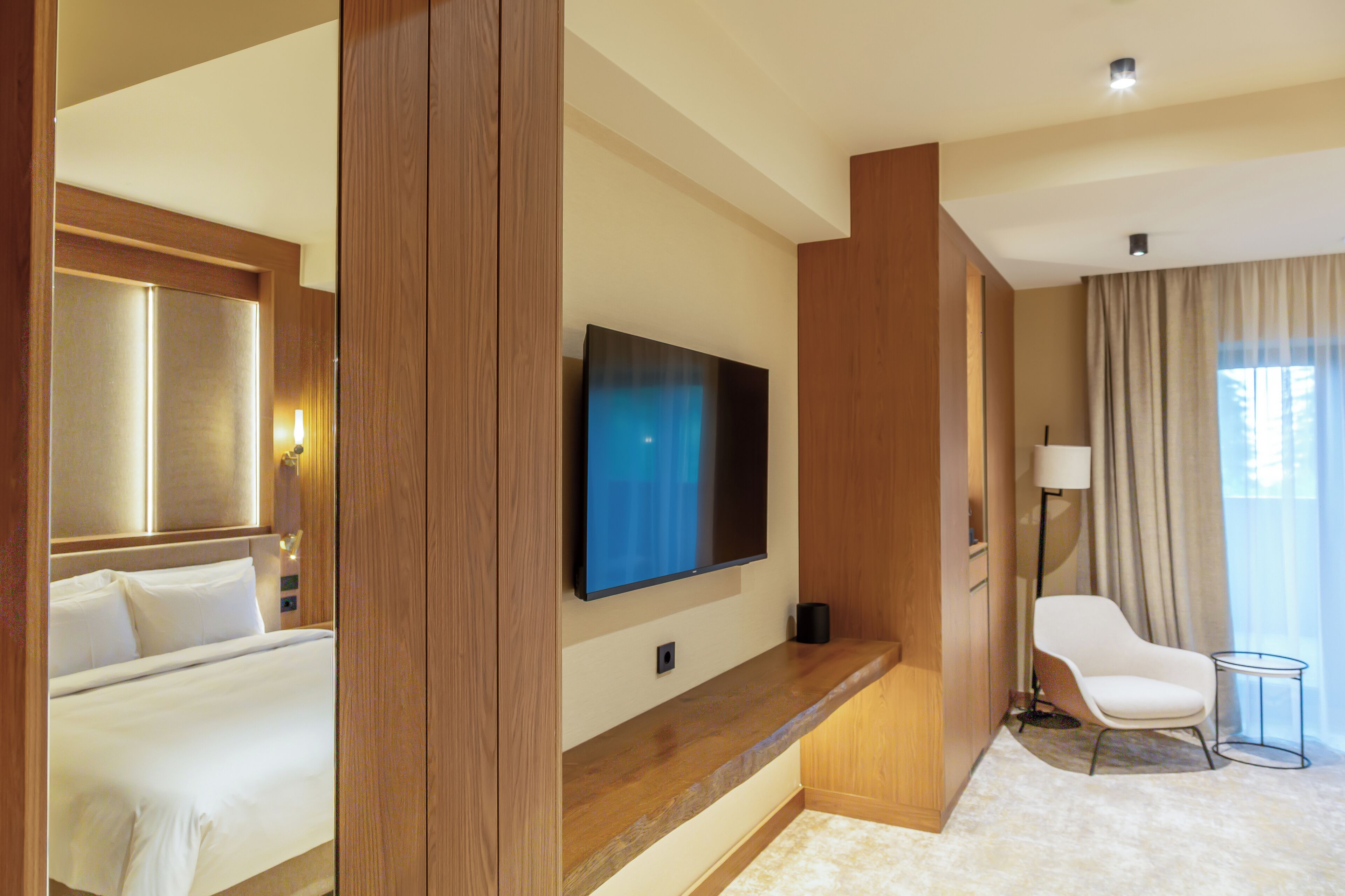 A hotel room with a bed, a flat-screen TV mounted on the wall, a seating area with a chair and a small table, and a large window with curtains.