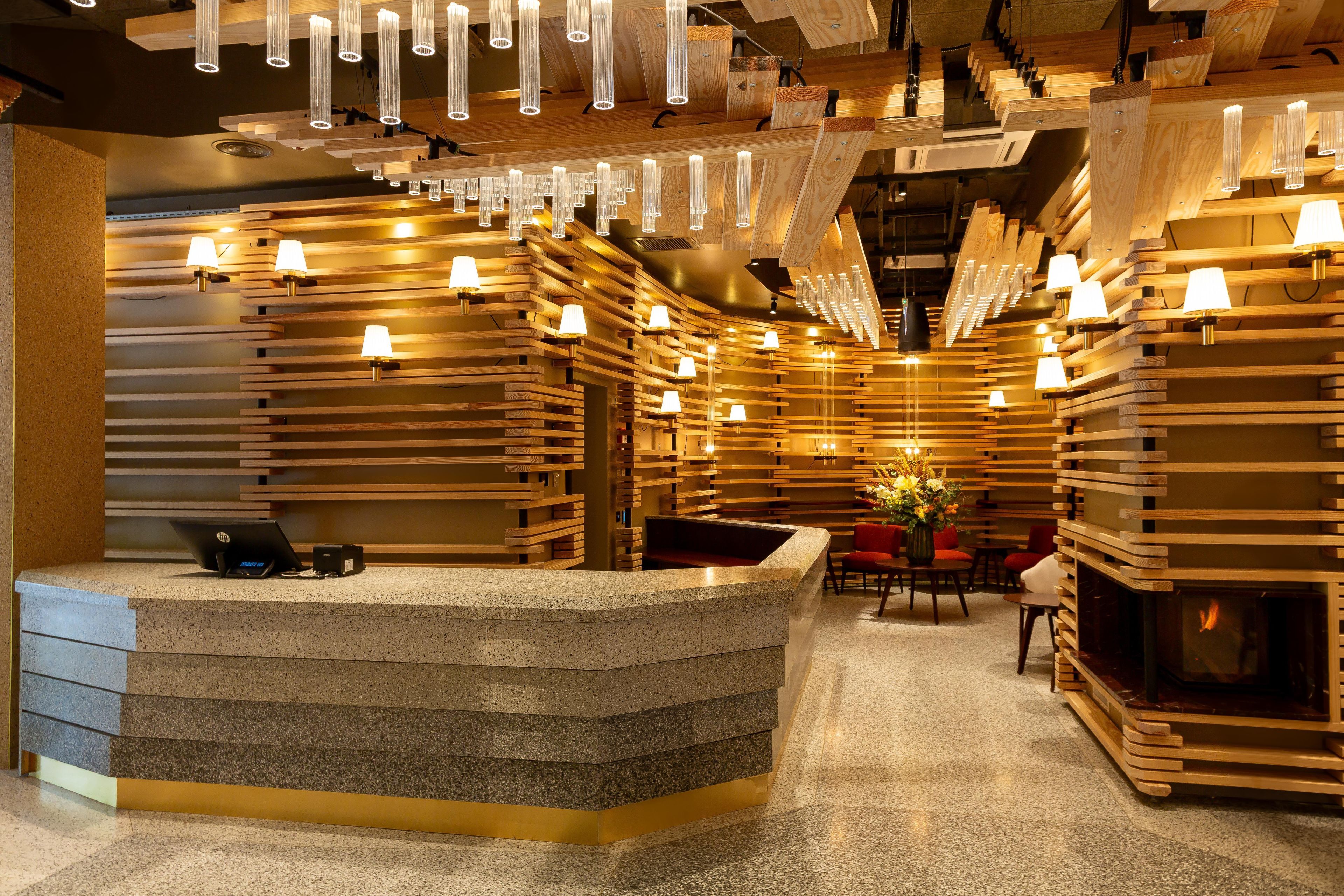 Un área de recepción con un mostrador de madera curvo, varias lámparas de pared y una zona de estar con sillas y una mesa.