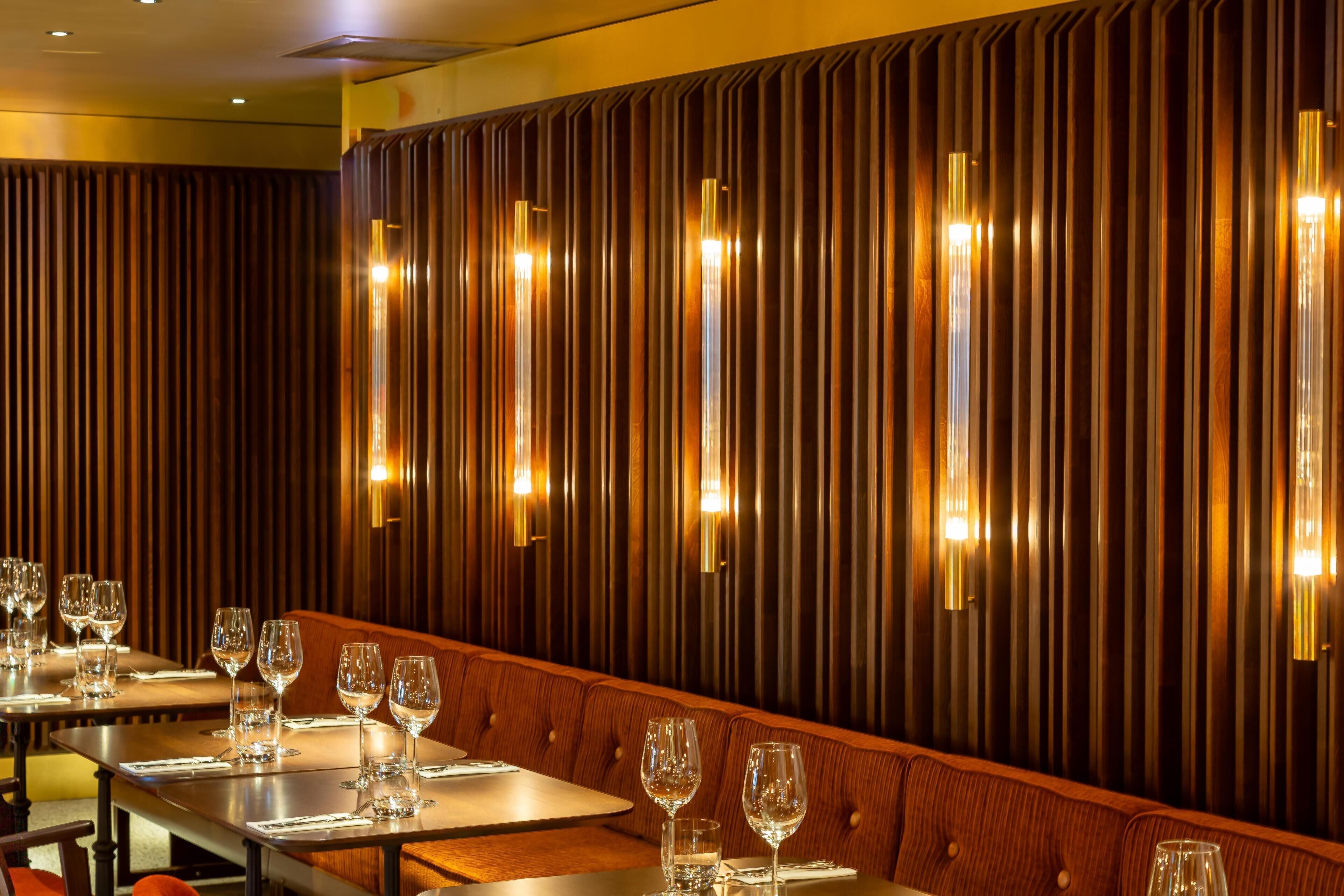 Interior de restaurante con una larga mesa de comedor, varias sillas y lámparas verticales en la pared.