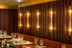 Interior de restaurante con una larga mesa de comedor, varias sillas y lámparas verticales en la pared.