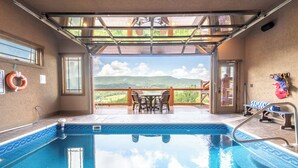 An indoor swimming pool with a view of a mountainous landscape, a covered patio area with a table and chairs, and a lifebuoy on the wall. - The Lodge at Coopers Hawk | Indoor Pool + Game Room & Hot Tub + VIEWS (Sevierville)