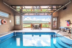 An indoor swimming pool with a view of a mountainous landscape, a covered patio area with a table and chairs, and a lifebuoy on the wall. - The Lodge at Coopers Hawk | Indoor Pool + Game Room & Hot Tub + VIEWS (Sevierville)