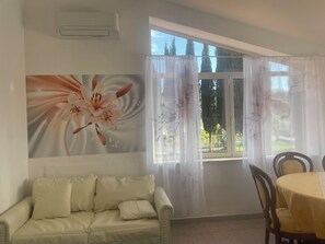 A room with a beige sofa, a dining table with chairs, a large window with sheer curtains, and an air conditioning unit on the wall. - Casa Maria della Rocca (Rocca d'Arce)