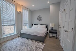 Une chambre à coucher comprenant un lit, une table de chevet, une lampe, un tableau ou une décoration murale, et une porte.