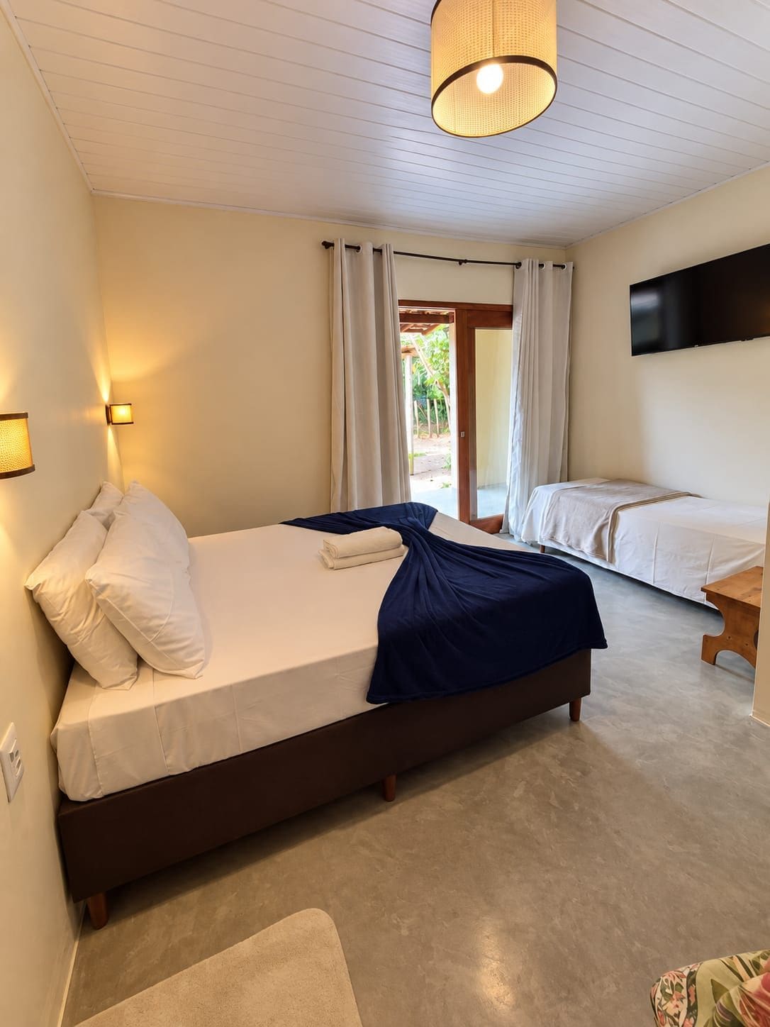 A hotel room with a large bed, a bedside table, a flat-screen TV mounted on the wall, and a view of the outdoors through a sliding door.