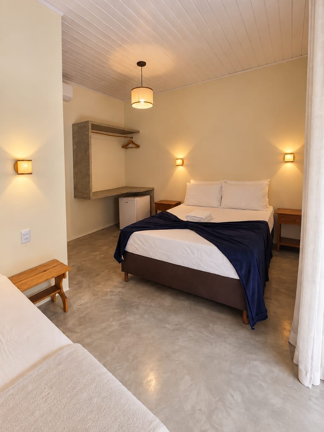Quarto com cama, mesas de cabeceira, luminária pendente e pequena escrivaninha com cadeira.