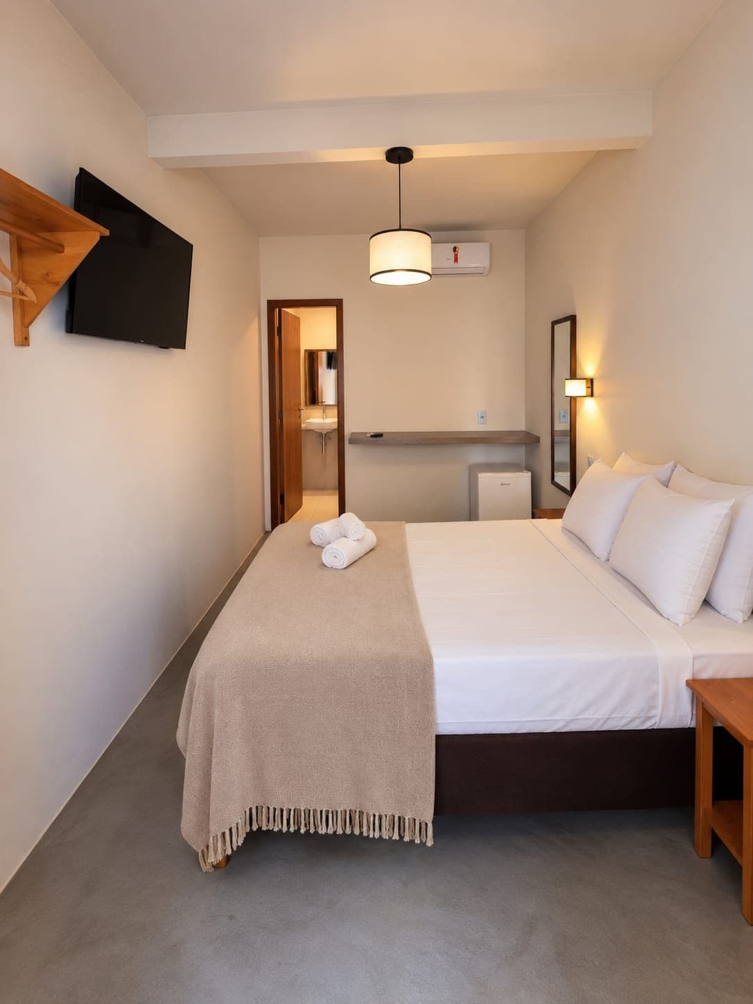 Quarto de hotel com cama, mesa de cabeceira, TV fixada na parede, espelho e luminária no teto.