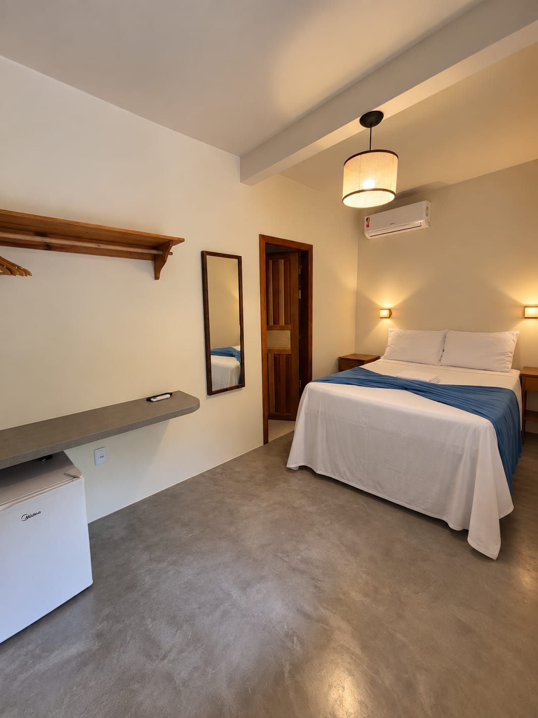 Quarto com cama, mesas de cabeceira, espelho, prateleira e um pequeno frigorífico.