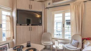 A modern room with a flat-screen TV, a dining table with plates and glasses, a chair with a pillow, a small side table, and a window with curtains. - Authentic Bosnian-Herzegovinian Studio & Garden in the heart of Mostar (Mostar)