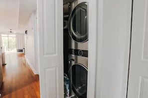 Une buanderie avec des machines à laver superposées et un couloir menant à une pièce lumineuse dotée d’un ventilateur de plafond.