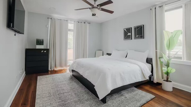 Ein modernes Schlafzimmer mit Bett, Deckenventilator, Fernseher, Kommode und einem Fenster mit Vorhängen.