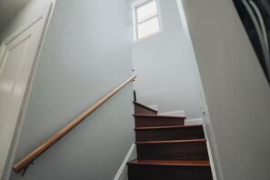 Una escalera con barandillas de madera y peldaños blancos, que lleva a un rellano con un ventanal.