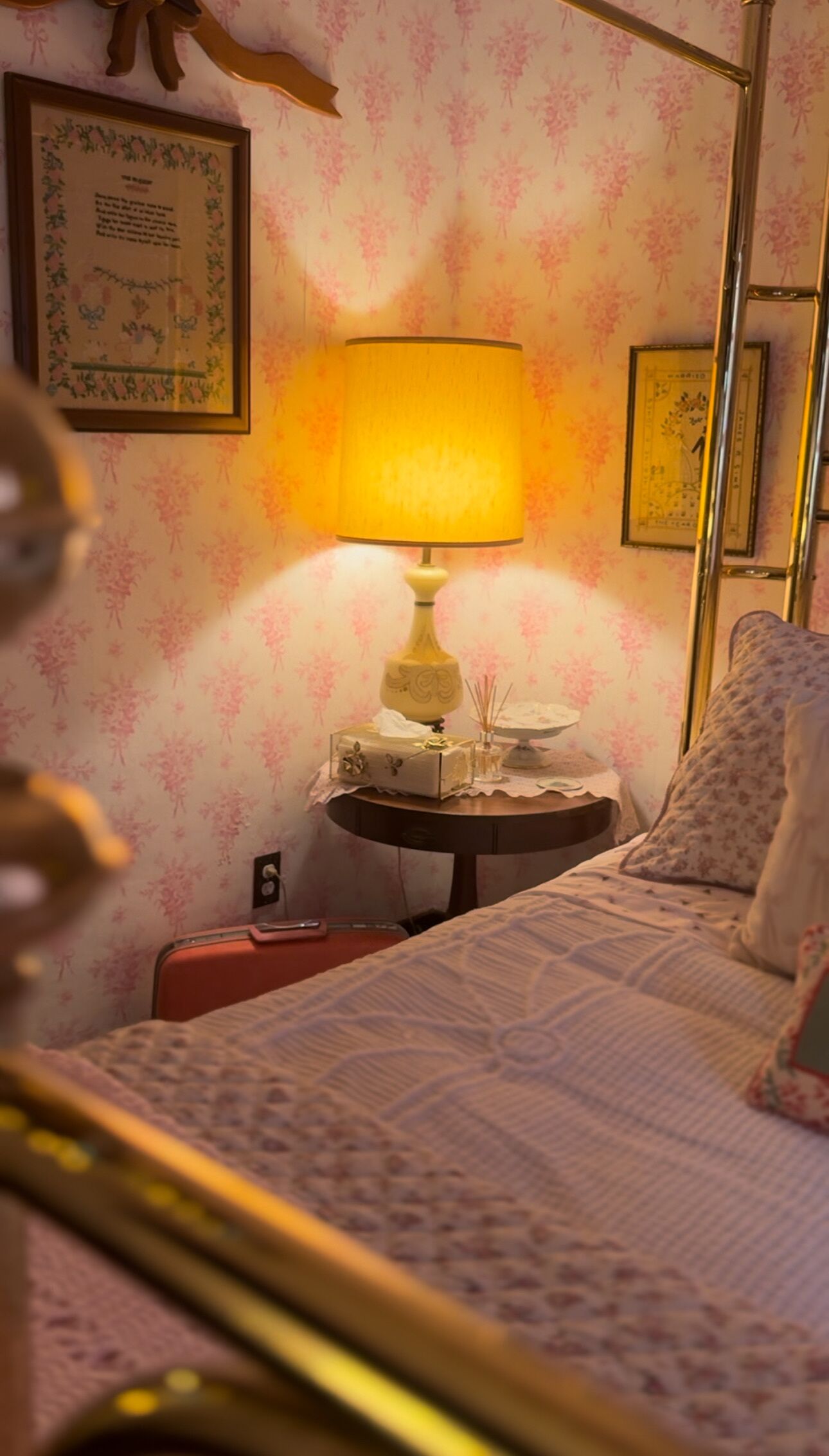 Un lit avec une couvre-lit à motifs, une table de chevet avec une lampe, un tableau encadré au mur et un papier peint à motifs floraux.
