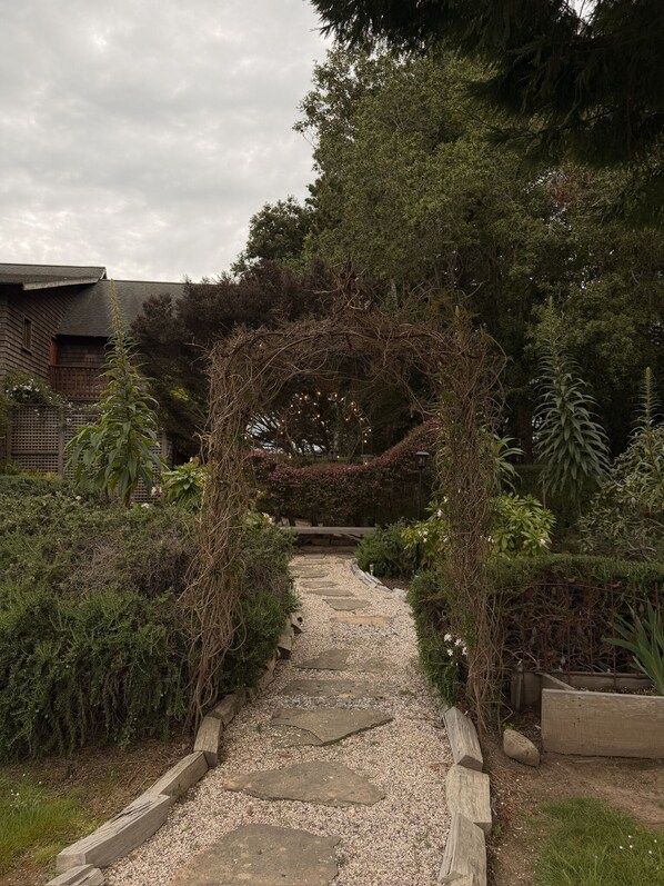 A stone pathway leading through a garden with a wooden archway, surrounded by various plants and trees.