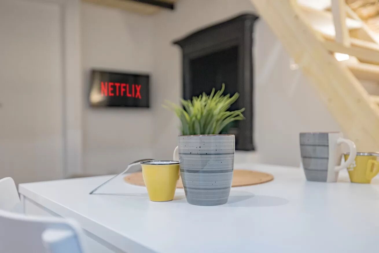 Una mesa blanca con una planta en maceta, una taza amarilla y una taza gris. Se ve un cartel con 'NETFLIX' en la pared.