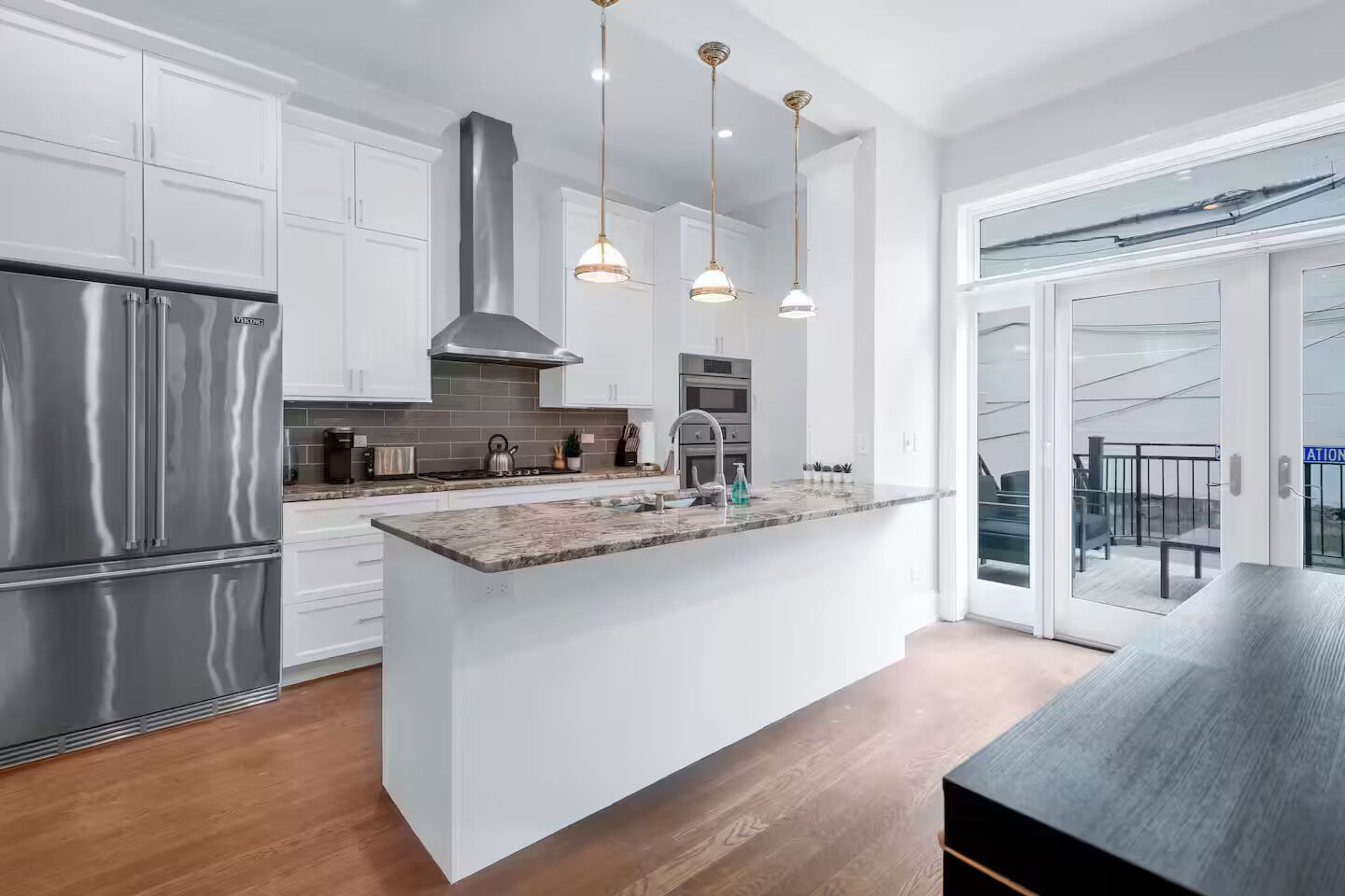 A modern kitchen with white cabinetry, stainless steel appliances, a central island, pendant lighting, and a view to a balcony.