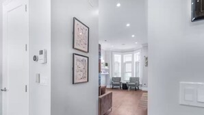 A hallway with white walls, two framed abstract artworks, a wooden bench, and a seating area with two chairs. - Gold Coast Pristine 5BR - 3.5BA - Patio - Parking (Chicago)