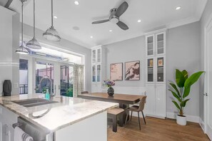 Una cocina moderna con una isla de mármol, luces colgantes, ventiladores de techo y un comedor con mesa y sillas.