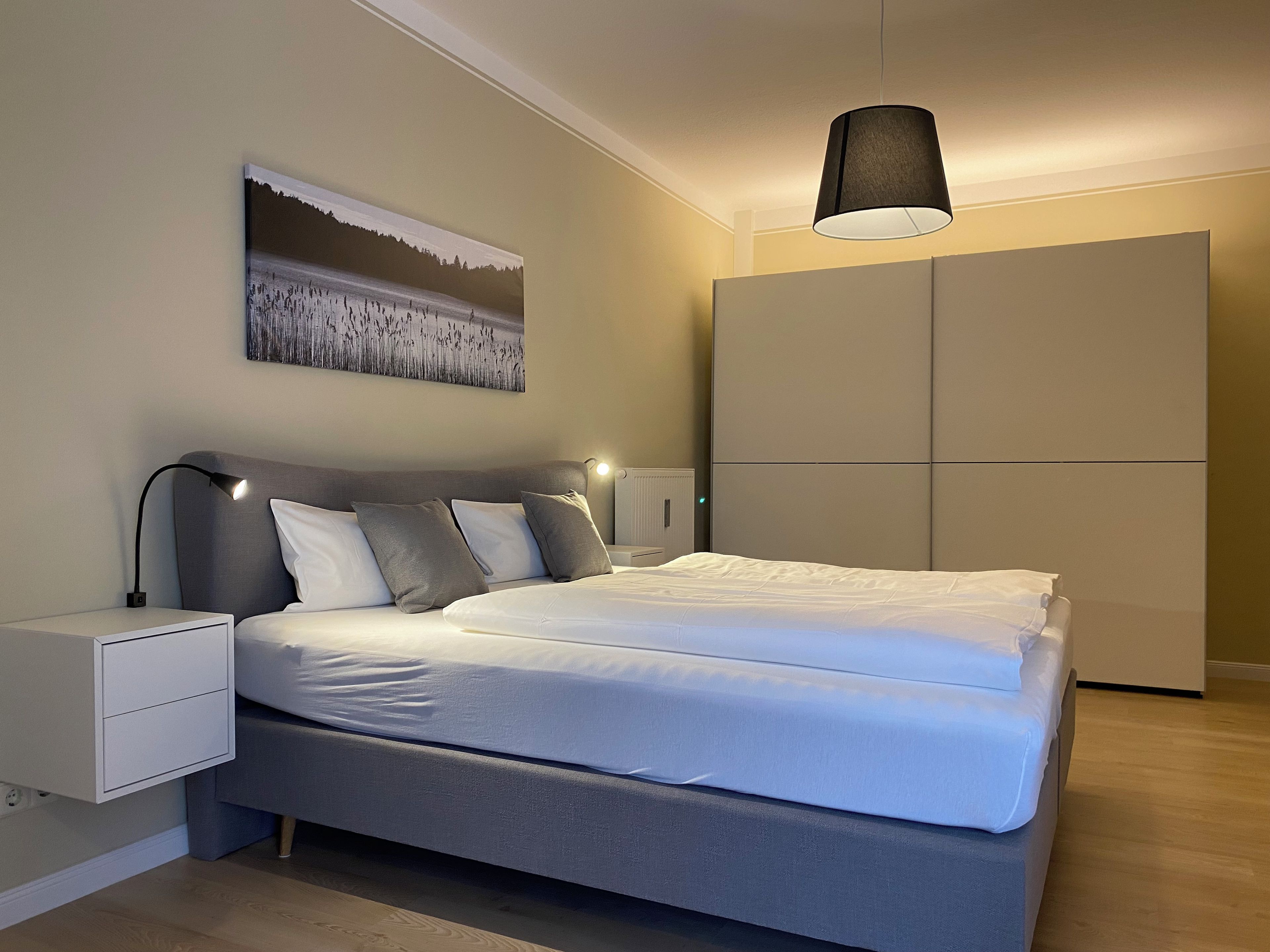 Ein modernes Schlafzimmer mit Bett, Nachttischen, einer Hängeleuchte, einem Kleiderschrank und einem Bild an der Wand.