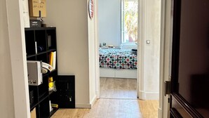 A hallway with wooden flooring, a black shelving unit, and a door leading to a room with a patterned bedspread.