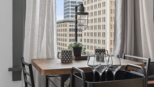 Un coin repas comprenant une table, des chaises, une plante en pot et une fenêtre donnant sur la ville.