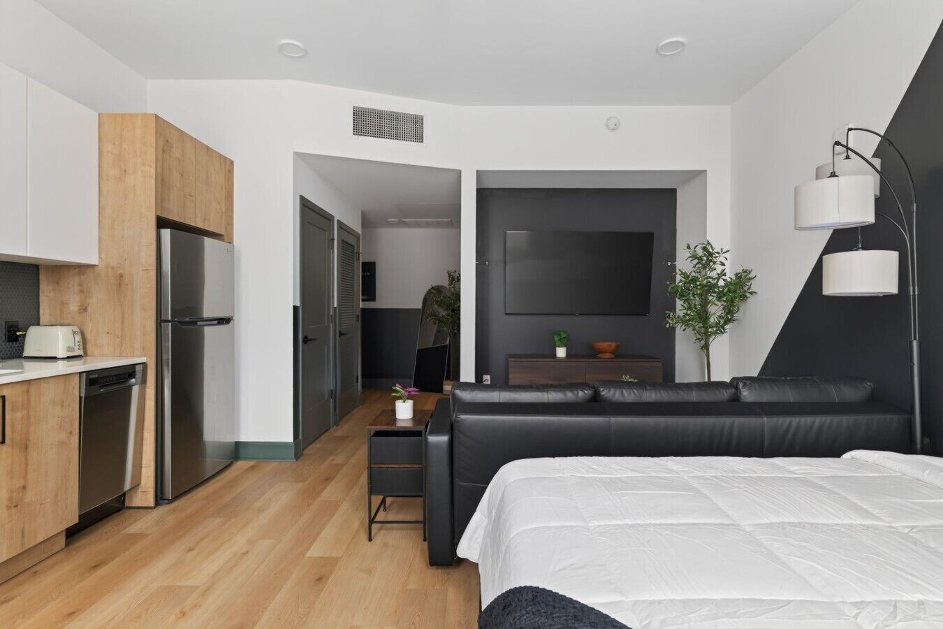 Une chambre d’hôtel moderne équipée d’une kitchenette, d’un grand lit, d’un canapé noir, d’une télévision et d’une plante.
