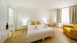 Une chambre d’hôtel avec un lit, des tables de chevet, un fauteuil jaune et une fenêtre avec des rideaux.