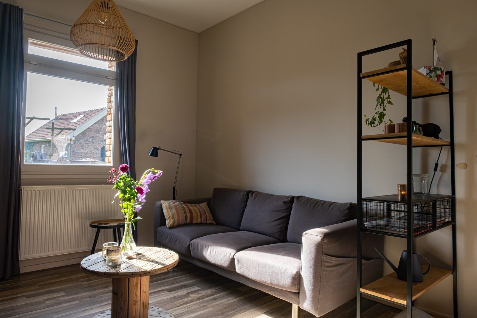 Ein Wohnzimmer mit einer Couch, einem runden Holztisch, einer Stehlampe, einem Bücherregal und einem Fenster mit Vorhängen.