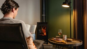 Una persona llegint al costat de la llar de foc, amb un llibre, un got de vi i un plat de menjar damunt d'una taula.