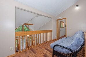 Ein Zimmer mit Holzboden, einem Bett mit grauer Matratze, einer Holzvertäfelung und Blick auf einen Balkon mit Holzgeländer.