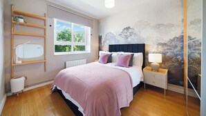 Ein Schlafzimmer mit einem Bett, einem Nachttisch, einer Lampe, einem Heizkörper, einem Fenster und einer Wand mit einem Wandbild von Naturmotiven.