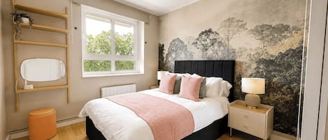 A bedroom with a bed, bedside table, lamp, window, and wall art.