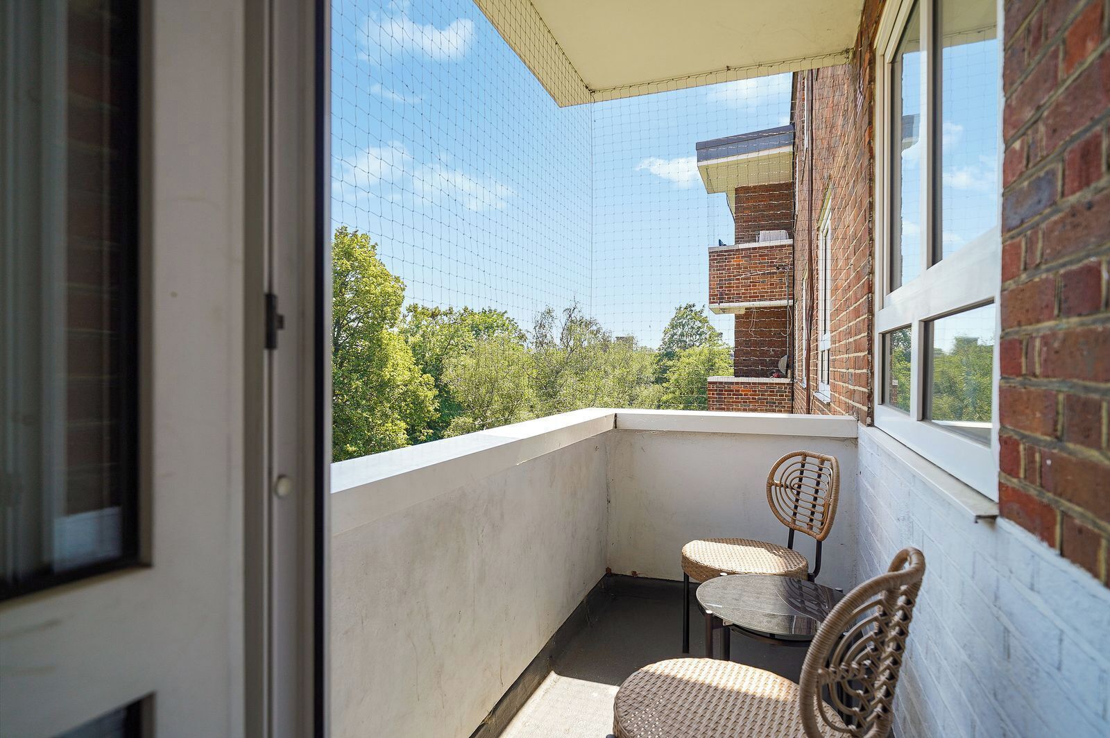 A balcony with a view of trees and a brick building. There are two wicker chairs and a small table.