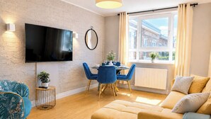 Ein modernes Wohnzimmer mit Flachbildfernseher, einem Essbereich mit blauen Stühlen, einer Couch und einem Fenster mit Vorhängen.